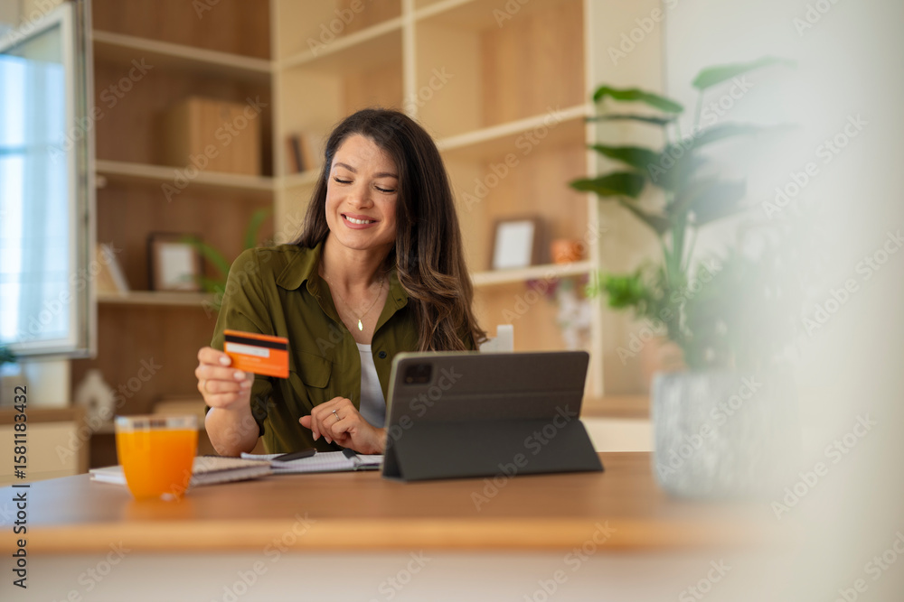 Fototapeta premium Woman with credit card using tablet at kitchen table, smiling and relaxed. 