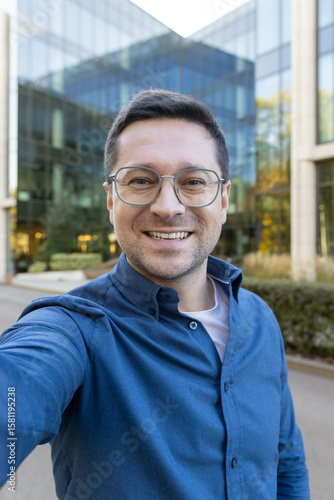 Konstfotografi A man wearing glasses takes a selfie in front of a modern glass building, smiling towards the camera
