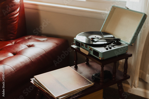 Vintage portable record player with open lid on small wooden stand beside red leather sofa with vinyl records in warm softly lit cozy living room interior