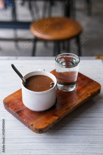 Steaming cup of kopi susu served with a glass of water on a rustic wooden tray at a cozy local coffee shop. Warung kopi Pontianak.