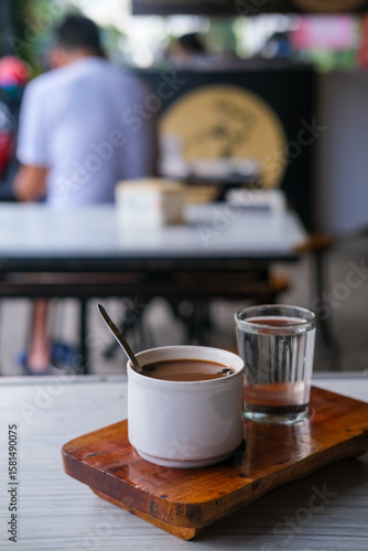 Steaming cup of kopi susu served with a glass of water on a rustic wooden tray at a cozy local coffee shop. Warung kopi Pontianak.