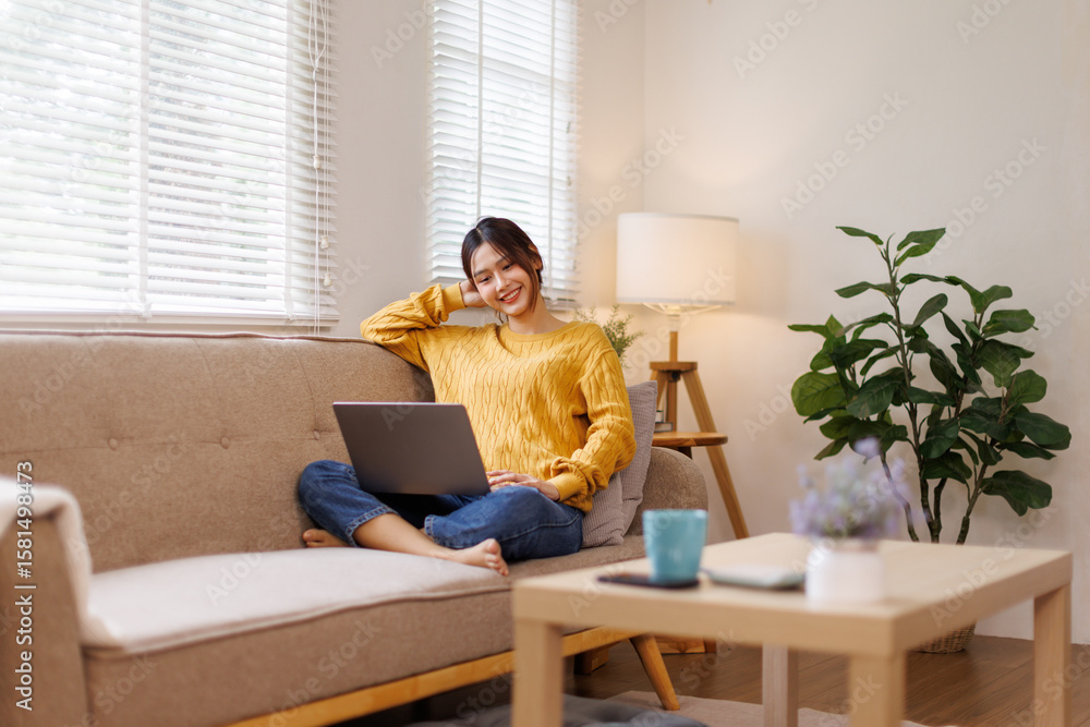 Young Asian woman doing research work for her business. Smiling woman sitting on sofa relaxing while browsing online shopping website. Happy girl browsing through the internet during free time at home Young Asian woman doing research work for her business. Smiling woman sitting on sofa relaxing while browsing online shopping website. Happy girl browsing through the internet during free time at home