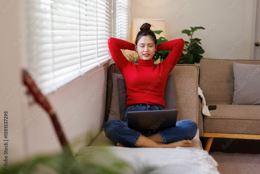 Obraz premium Rest Concept. Happy asian young woman sitting on comfortable couch at home in living room. Cheerful casual woman relaxing on sofa, leaning back, enjoying weekend free time or break from work 