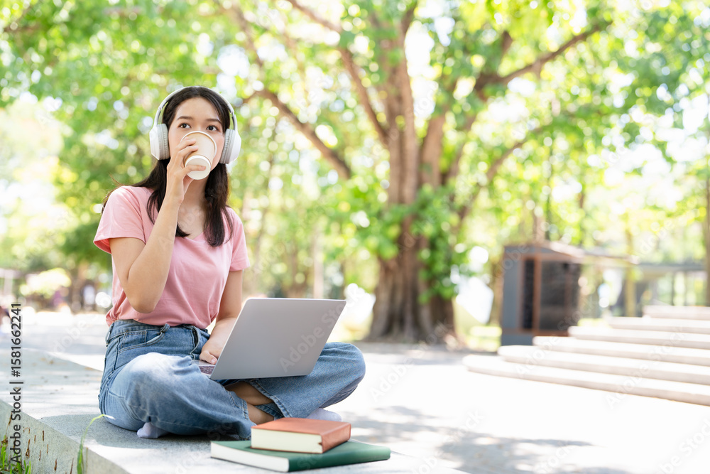 Young female student with laptop and headphones studying online sitting outdoor drinking coffee at university campus. Young female student with laptop and headphones studying online sitting outdoor drinking coffee at university campus.