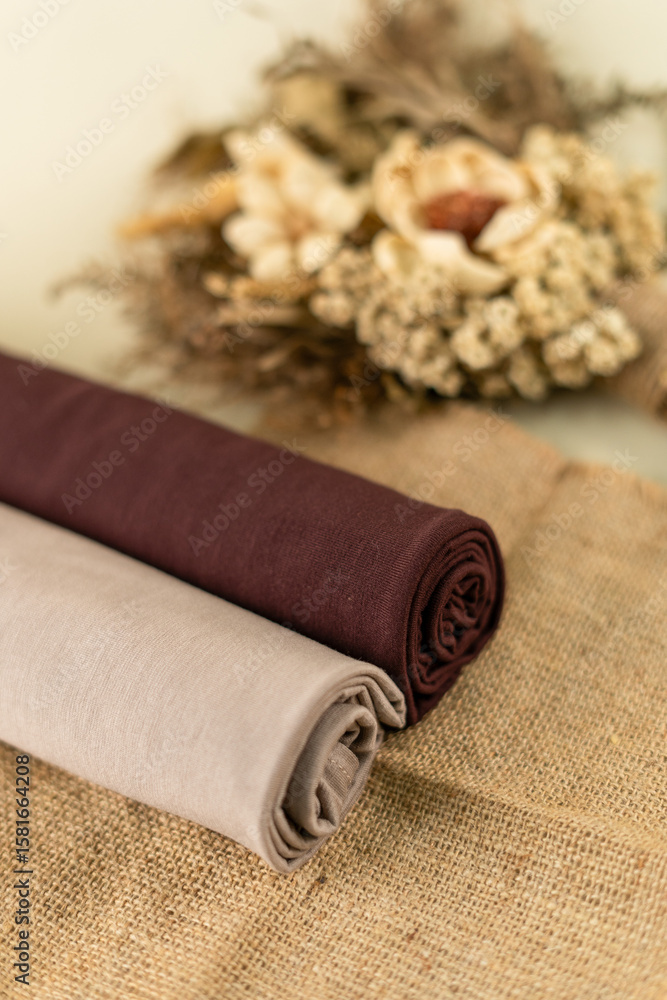 Fototapeta premium Two Rolled Pashmina Hijabs In Beige And Brown With Dried Flower Decoration On A Burlap Fabric For Fashion Product Display, Shot In Soft Natural Lighting With A Minimalist Style.
