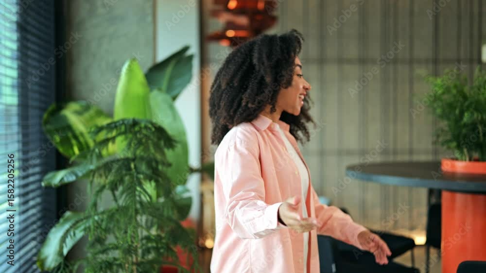 Confident African American businesswoman with curly hair, dressed in a pink shirt and white t-shirt, stands smiling in a bright modern office, gesturing invitingly. Positive and cheerful emotions. Confident African American businesswoman with curly hair, dressed in a pink shirt and white t-shirt, stands smiling in a bright modern office, gesturing invitingly. Positive and cheerful emotions.