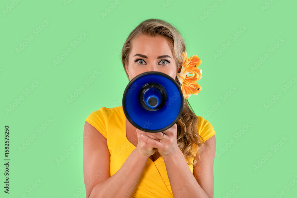 Fototapeta premium Woman in yellow shirt using blue megaphone with flower in hair promoting event against green background