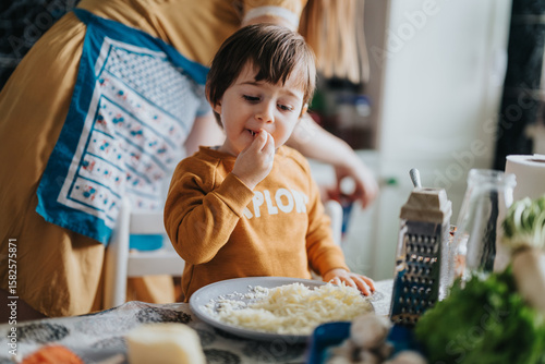 A child wearing a yellow shirt eats grated cheese from a plate while cooking alongside an adult in a bright and pleasant kitchen environment.