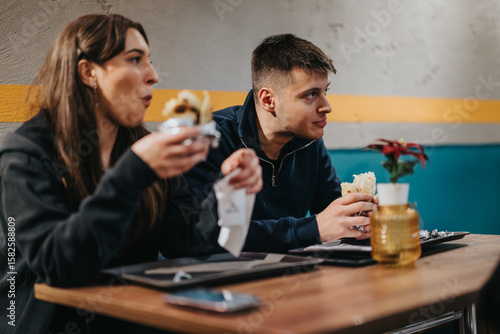 Two young people savor their meals indoors, displaying friendship and enjoyment in a comfortable setting.