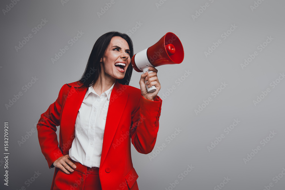 Confident young woman in a red suit using a megaphone to communicate passionately in a professional setting on gray background Confident young woman in a red suit using a megaphone to communicate passionately in a professional setting on gray background