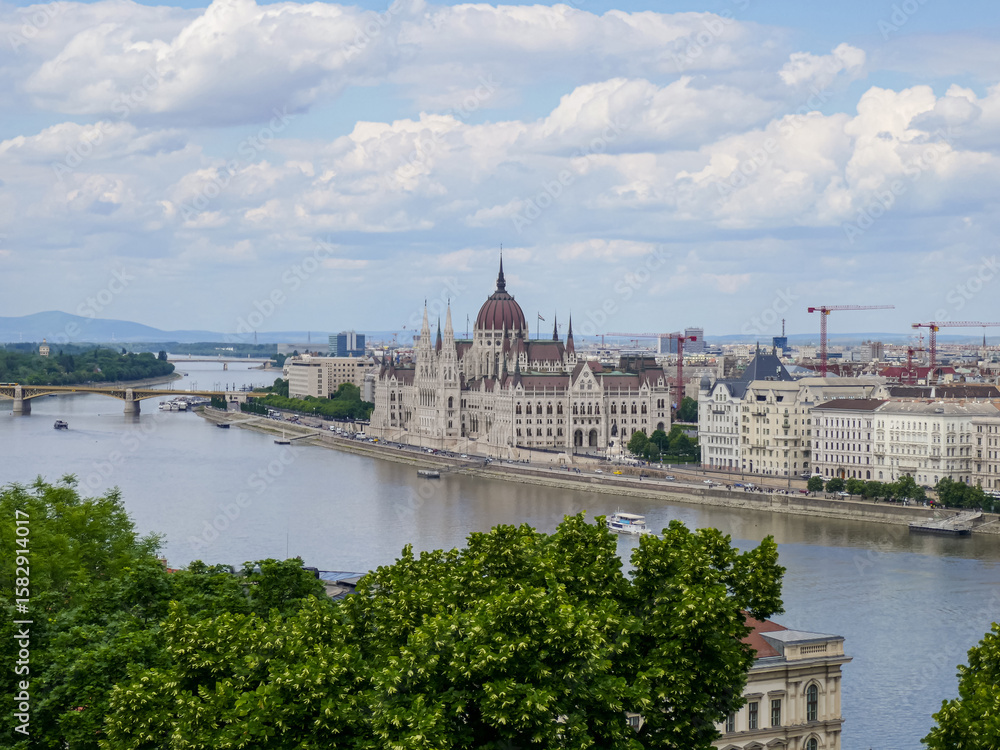 Fototapeta premium Scenic view of Hungarian Parliament Building in Budapest framed by green trees and cloudy sky from across the river.