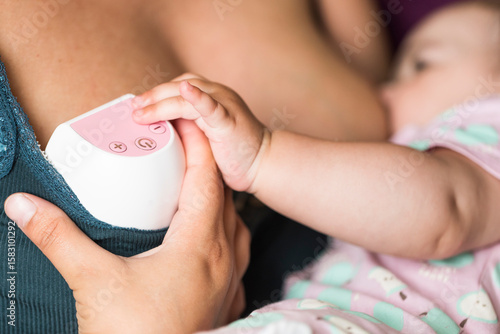 Mother using a wearable electric breast pump while breastfeeding her baby. Real moment of modern lactation, milk expression, and maternal care at home