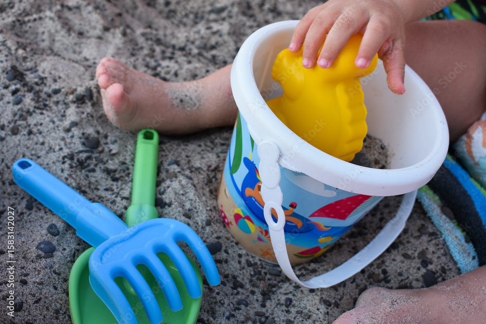 Little kid sitting on the towel and playing with plastic toys. Little girl playing with plastic bucket, shovel, spade and mold on a sandy beach. Summer vacation in Sicily, Italy. Little kid sitting on the towel and playing with plastic toys. Little girl playing with plastic bucket, shovel, spade and mold on a sandy beach. Summer vacation in Sicily, Italy.