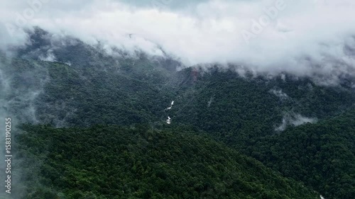 An epic drone view of a cascading waterfall lush mountains and forests topped with misty clouds