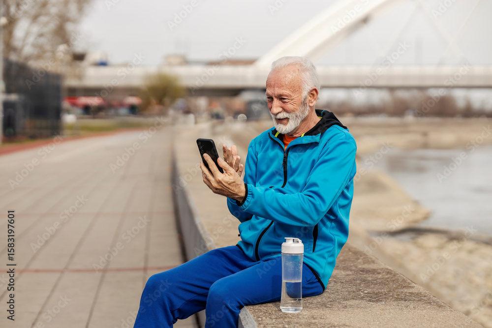 Fototapeta premium Portrait of senior sportsman sitting on riverside and having online video call.