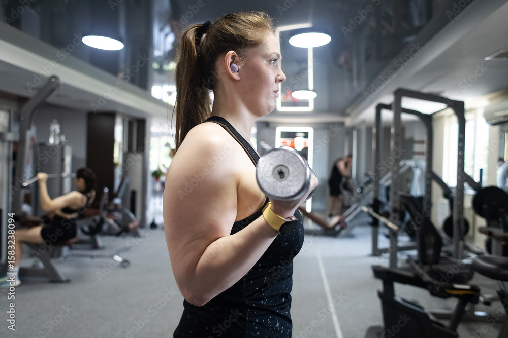 Fototapeta premium Ordinary woman in the gym doing exercises with dumbbells. Workout for weight loss. Body strengthening and healthy lifestyle
