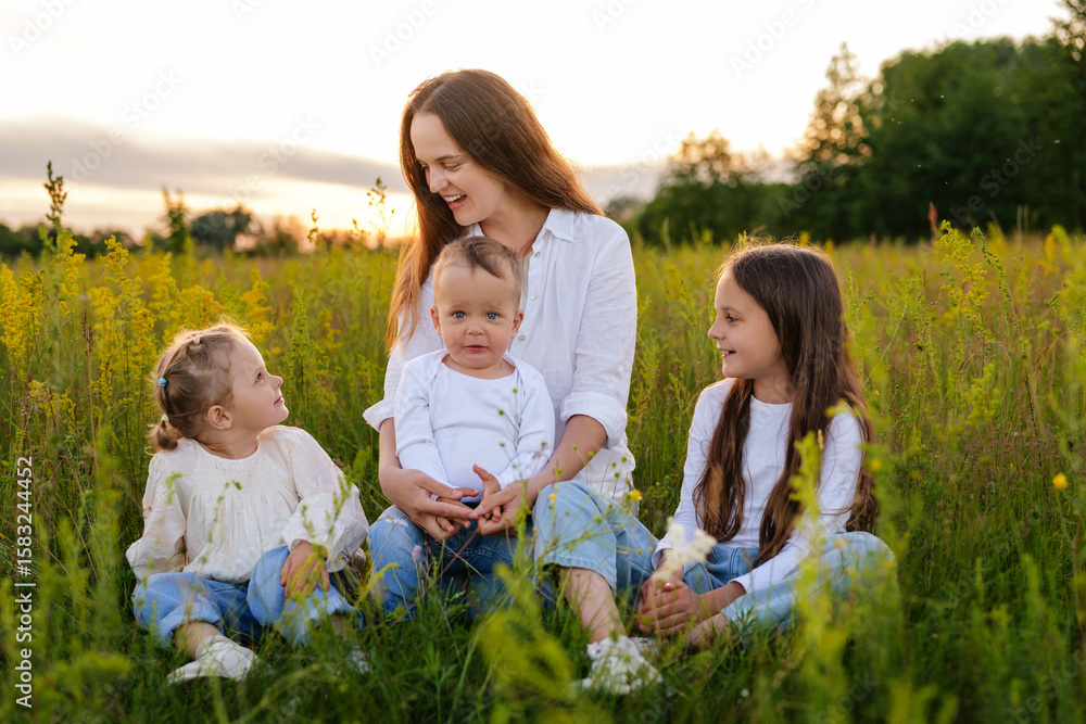 A joyful mother sits in a sunlit grassy field surrounded by her three children (two girls and a baby), all wearing white tops and denim, enjoying the outdoor evening. A joyful mother sits in a sunlit grassy field surrounded by her three children (two girls and a baby), all wearing white tops and denim, enjoying the outdoor evening.