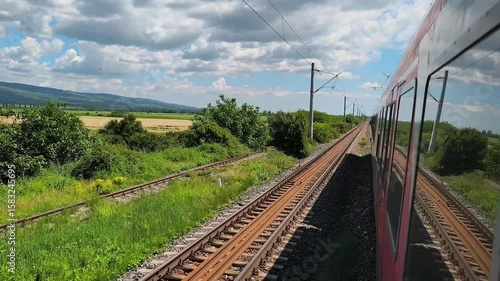 A red passenger train moves along a double-track railway through wide rural fields under a partly cloudy summer sky , in Romania