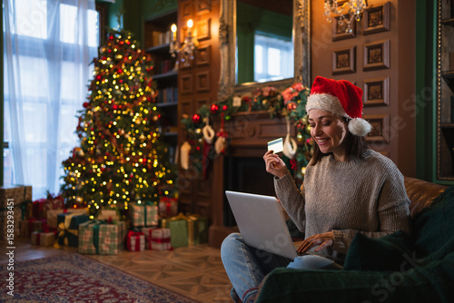 Merry Christmas. Young woman shopping online with laptop paying with gold credit card for Christmas gifts. Girl buying on Internet enter credit card details enjoying winter holidays at home