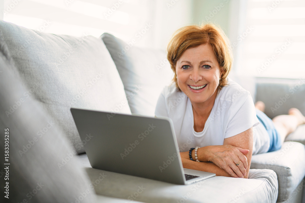 Happy modern European senior woman using laptop, touching eyeglasses and smiling to camera, enjoying digital leisure, sitting on couch Happy modern European senior woman using laptop, touching eyeglasses and smiling to camera, enjoying digital leisure, sitting on couch