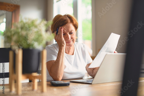 Sad frustrated senior woman pensioner having depressed look, holding hand on her face, calculating family budget, sitting at kitchen counter with laptop, papers, coffee, calculator and cell phone