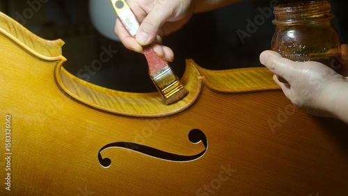 Fotografie Skilled luthier applying varnish on cello body, holding stained jar, highlightin