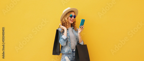 Beautiful happy young woman holding phone with black shopping bag, summer hat on yellow background