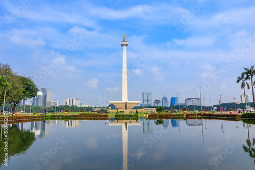 National Monument (Monas) is iconic landmark in the center of Jakarta reflected in a calm pool with palm trees and blue sky.