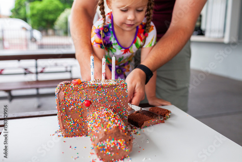 Three year old girl cutting birthday cake shaped like a camera with hundreds and thousands