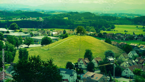 Lonely Tree on a Hill Overlooking a European Town – Scenic Landscape. A scenic panoramic view of a small European town nestled in lush greenery, with a single tree standing atop a grassy hill. 
