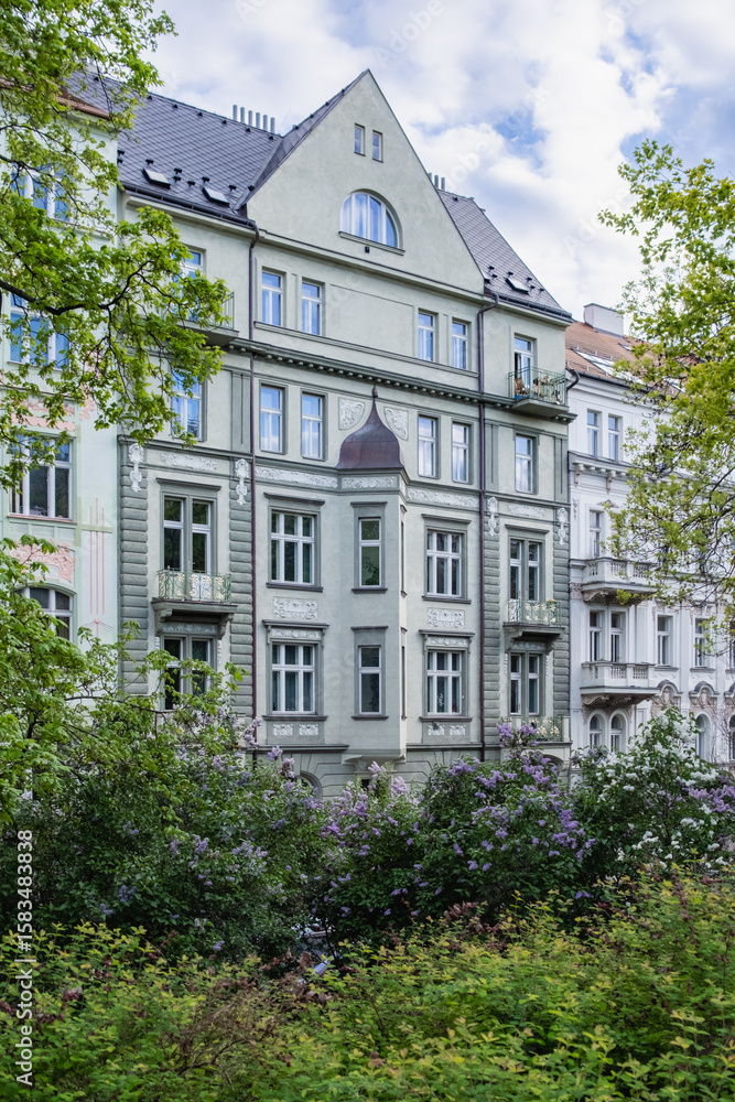 Fototapeta premium Elegant historic residential building in Prague, Czech Republic, framed by blooming lilac bushes and spring greenery, showcasing beautiful European architecture and ornate window details.