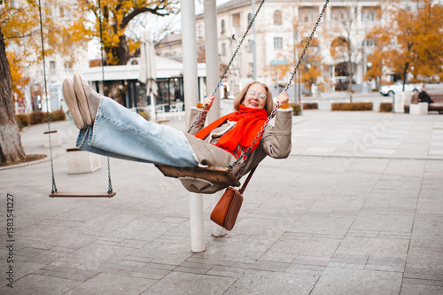 Smiling mature senior woman 55-59 year old rocking on swing at city street wearing vision glasses outdoor. Senior adult lifestyle.