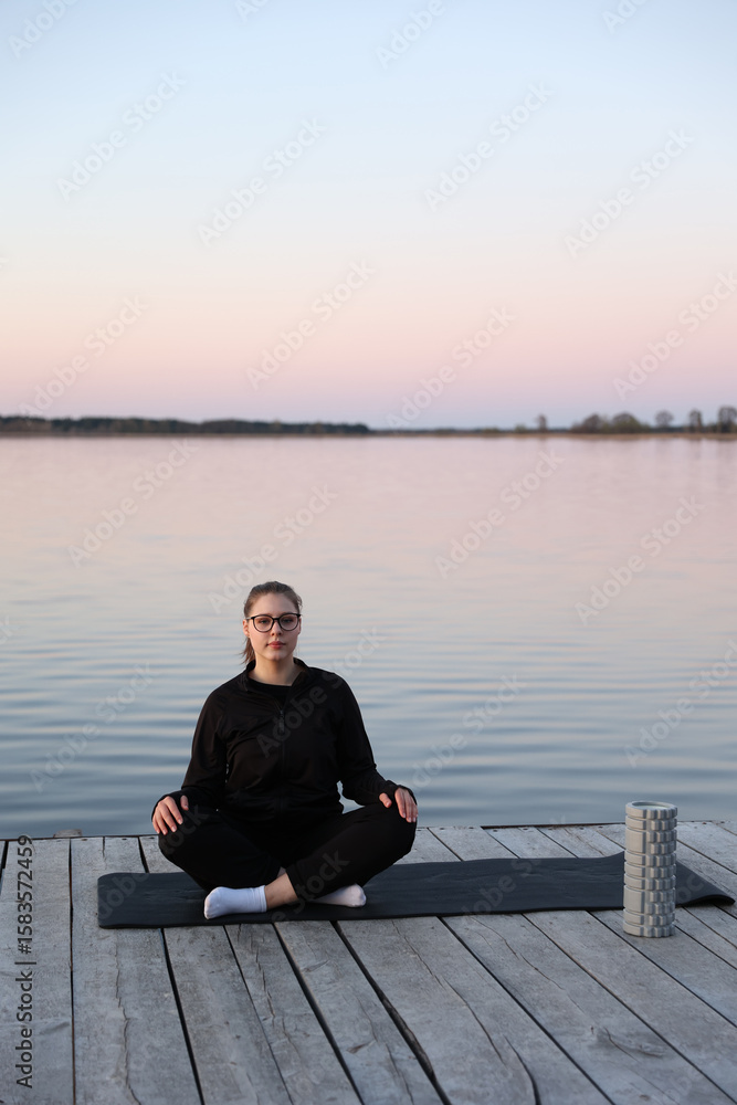 Fototapeta premium Woman practicing yoga on a wooden pier by the water at sunrise. Peaceful atmosphere, healthy lifestyle, balance and harmony with nature. Woman meditating on a yoga mat at wooden pier by the water