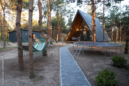 Modern A-frame cabin in a pine forest with hammock, trampoline, and string lights. Peaceful woodland retreat with cozy outdoor vibes