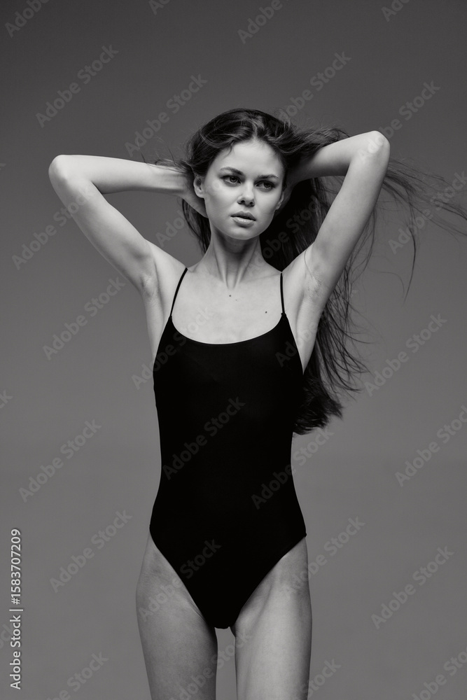 Fototapeta premium black and white woman posing in black swimsuit with hands behind head, long hair flowing, serious expression on gray background, studio lighting