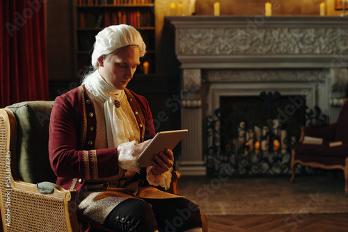 Fototapeta Caucasian young adult man with powdered wig and period costume sitting in ornate