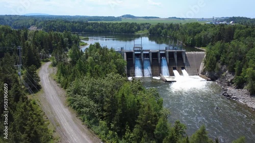 A view of a hydroelectric dam with cascading water and full-flow spillway gates, combining industrial design with a lush forest setting, showcases sustainable renewable energy production. Mitis-2 Dam.