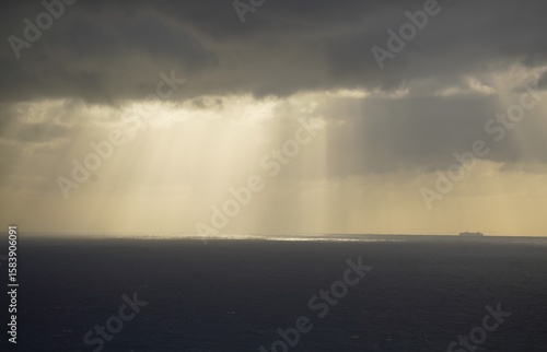 sun shines through distant clouds over the ocean , with ship in the rays of light