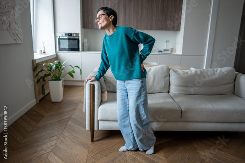 Mature woman hunched over in discomfort attempting to stand from sofa. Sciatica pain, slipped disc, arthritic inflammation, lower back injury, limited mobility, physical struggle, health limitation