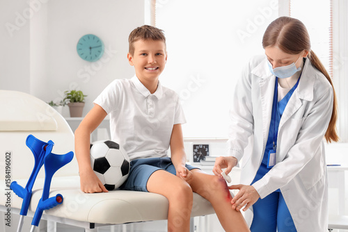 Smiling little boy with wounded knee and soccer ball visiting doctor in clinic