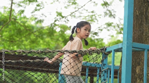 Happy young girl with pigtails waving cheerfully while standing on a net structure in a lush green outdoor setting surrounded by nature
