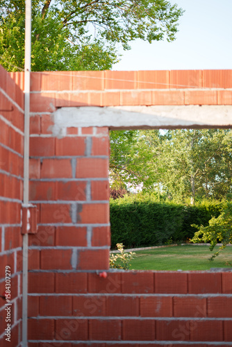 Red clay wall under construction with window opening framing the garden, symbolizing the connection between home and nature.