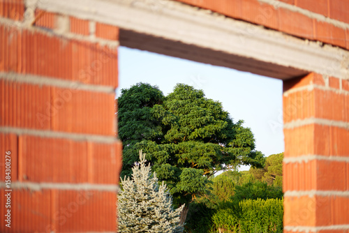 Red clay wall with open window framing green trees, symbolizing connection between sustainable home and natural surroundings.
