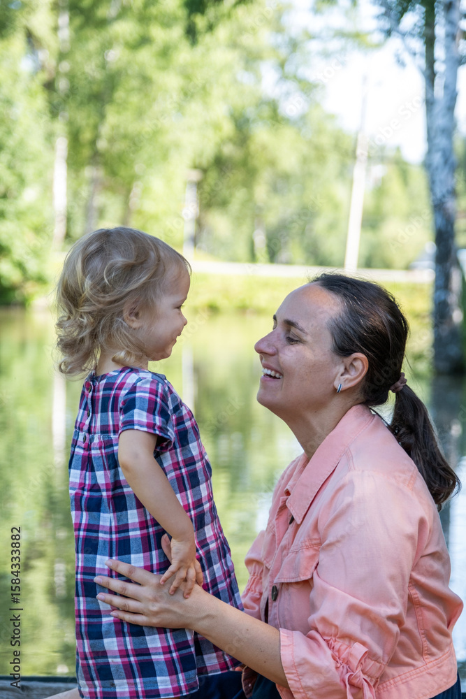 Fototapeta premium A joyful mom kneels to interact with her smiling daughter at a serene lake park. The sun shines brightly, creating a perfect summer ambiance and memories.