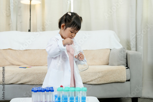 Little Asian girl wearing white lab coat experimenting mixing liquids using pipette in colorful test tubes playful science education activity at home bright cozy living room early learning