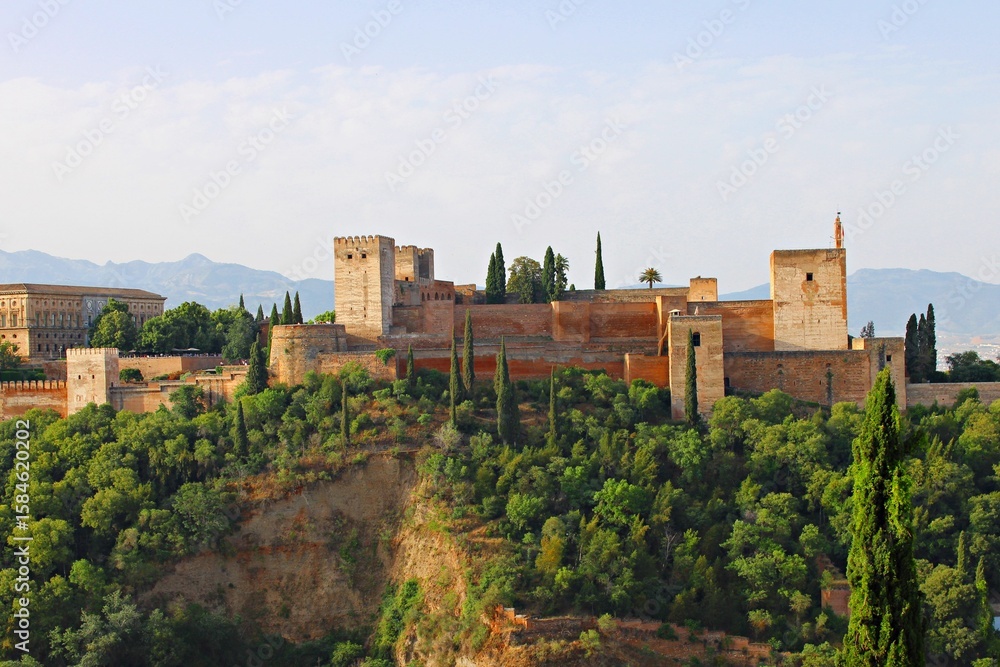Fototapeta premium Panoramic view of the arabic fortress Alhambra in Granada from the Mirador de San Nicolás, showcasing the Nasrid palaces, towers, and ancient defensive walls rising above the lush forested hillside.