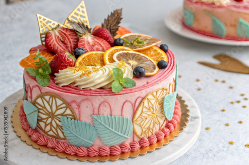  shows a beautifully decorated cake with a pink buttercream frosting, adorned with various fruits like strawberries, blueberries, and orange slices, along with gold leaf decorations. The cake is place