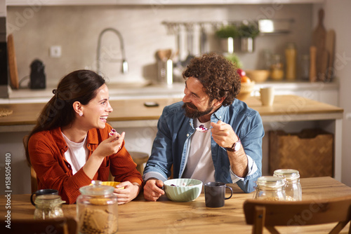 Photography Two friends share a light meal at a wooden dining table, smiling and engaged in conversation amidst a warm, well-lit kitchen filled with homey details