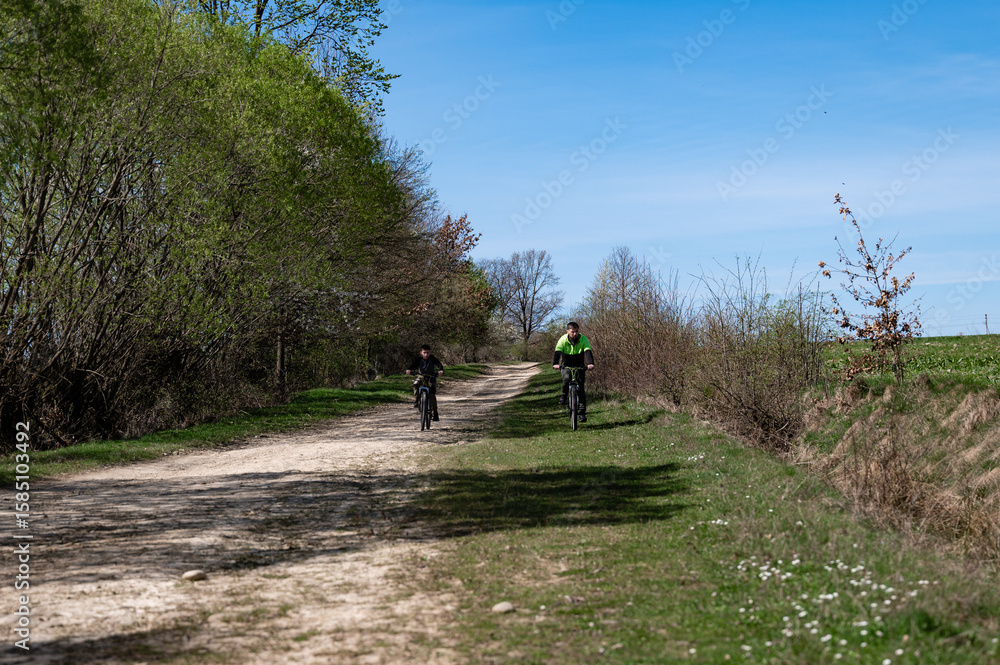 Obraz premium Two cyclists enjoy a sunny ride down a country lane, surrounded by lush greenery.