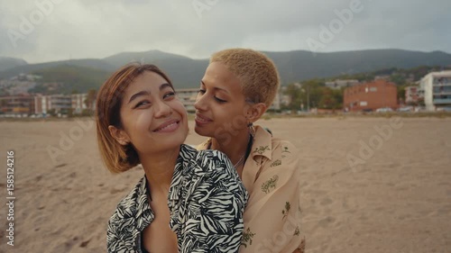 Passionate lesbian couple sharing tender moment while embracing and kissing on sandy beach during romantic sunset, expressing deep love and connection